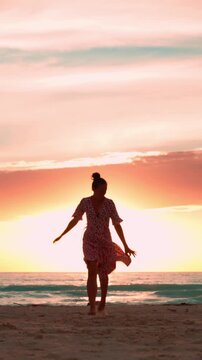 Silhouette of a happy woman in a summer dress running barefoot on the sandy beach at sunset, enjoying her freedom and happiness during a beautiful summer vacation. Vertical video