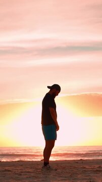 Young bearded man feeling lonely standing on a sandy beach during a beautiful warm sunset, contemplating the ocean, feeling emotional and introspective during travel. Vertical video