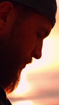 Close-up of sad thoughtful bearded man with sun flare at golden hour, contemplating life against a beautiful sunset over the sea, expressing solitude and sorrow. Vertical video