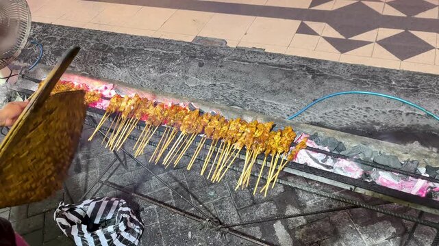 Mouthwatering street food moment as a vendor fans rows of Indonesian satay skewers grilling over glowing hot charcoal embers