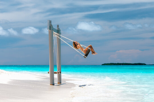 Young woman in light blue bikini swinging on beach swing above turquoise ocean water at tropical island resort