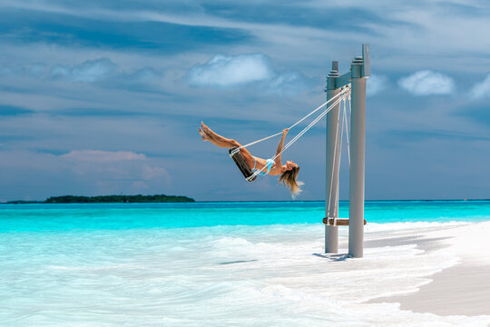 Young woman in light blue bikini swinging on beach swing above turquoise ocean water at tropical island resort