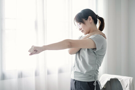 Young Asian woman stretching arm to relieve shoulder muscle pain near window inside office after long hours working for ergonomics or healthy lifestyle concept