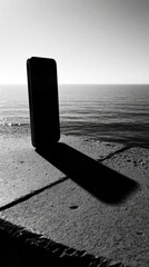 Fototapeta premium Black, white silhouette of smartphone on stone pier. Mobile device stands upright against ocean horizon background. Calm sea waves meet sky at coast. Minimalist tech concept with copy space for