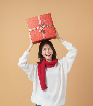 Cheerful Asian woman wearing a white sweater and carrying a red gift box on the shoulder while smiling.