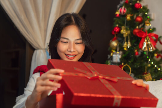 Cheerful Asian woman sitting on a sofa, excitedly opening a large red gift box at home with Christmas decorations in the background, holiday celebration.