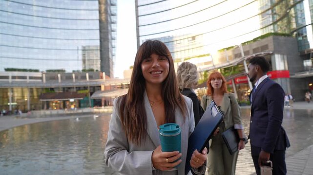 Portrait of a successful businesswoman smiling at the camera, holding a reusable coffee cup and a folder with her multicultural colleagues talking in the background in a modern city downtown