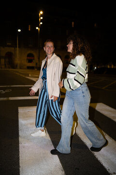 Two white women walking on a crosswalk together at night