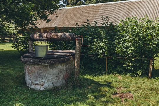 Old rustic stone water well with metal bucket in sunny green garden. Rural atmosphere, charm of countryside living. Nostalgia, simplicity, tradition, self-sufficiency in natural environment.