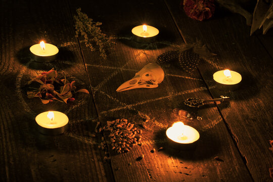 Witchy altar with drawn pentagram circle decorated burning candles, a bird skull, dried herbs, autumn flowers, crystals and an old key on wooden surface in the dark