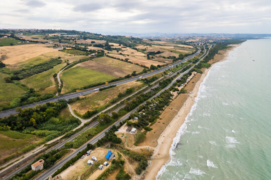 Aerial view of the Molise coast in Italy, showing a sandy beach along the turquoise Adriatic Sea. A coastal road, railway, highway, and seaside campsite border rolling agricultural fields and