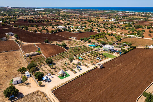 A high aerial view captures the rural Puglia countryside near Polignano a Mare, showcasing distinct red soil fields, scattered olive trees, and stone walls stretching towards the deep blue Adriatic