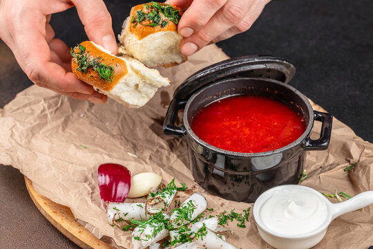 Hands dipping herb topped bread into bright red soup with onion garlic dill sour cream and rustic wooden board food scene