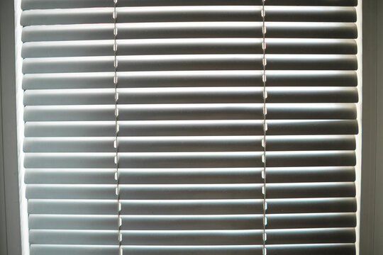 Horizontal gray blinds fill the window, with evenly spaced slats and hanging cords creating striped shadows across the indoor residential window covering