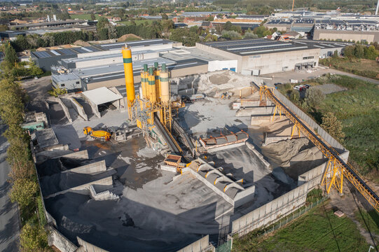 Aerial view of a concrete batching plant with yellow silos, conveyor belts, and piles of gravel in an industrial area of Roeselare, Flanders, Belgium.