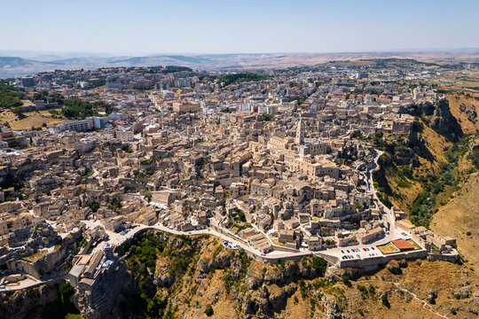 A high aerial panorama of Matera, Italy, showing the famous ancient Sassi quarter built along the edge of a dramatic canyon. The dense stone architecture, cathedral tower, and surrounding rugged