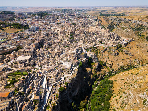 A high aerial panorama of Matera, Italy, showing the famous ancient Sassi quarter built along the edge of a dramatic canyon. The dense stone architecture, cathedral tower, and surrounding rugged