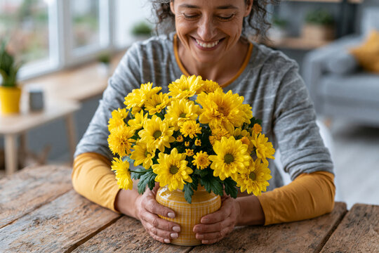 High quality composition of smiling adult arranging fresh flowers in a ceramic vase on a rustic wooden table during a diy home decor project, bright natural window light, cozy modern home interior, ,