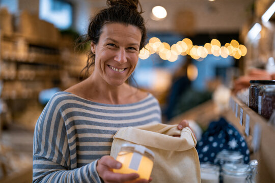 Smiling shopping with reusable produce bags and glass jars at a zero waste grocery store, clean composition
