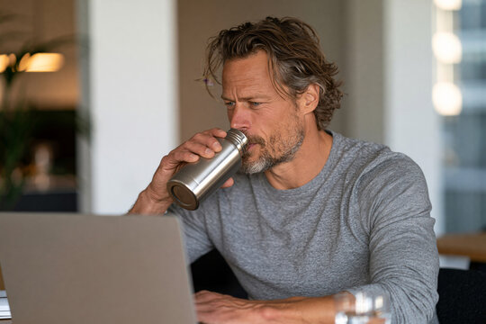 High quality composition of young professional drinking from a reusable stainless steel water bottle while working at a modern minimalist desk, clean workspace, natural daylight