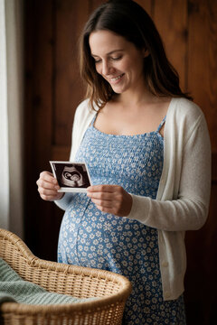 Smiling pregnant woman holds ultrasound photo of unborn baby. Happy expectant mother looks at sonogram in home interior. Caucasian female wearing blue floral dress, white cardigan feels joy waiting