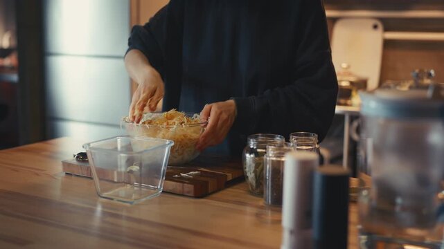 Woman hand mixing shredded cabbage and carrots with spices in a glass bowl. Preparing homemade fermented sauerkraut on a wooden table in a cozy kitchen. Close-up process of salting vegetables.