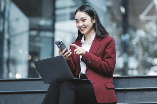 Focused woman in blazer types on laptop outdoors. Urban professional works remotely on stairs. She uses modern tech for business and connection.