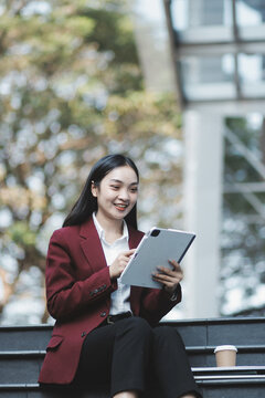 Focused woman in blazer types on laptop outdoors. Urban professional works remotely on stairs. She uses modern tech for business and connection.