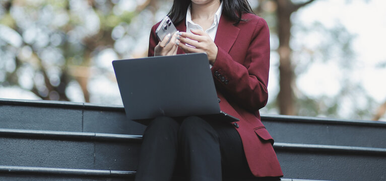 Focused woman in blazer types on laptop outdoors. Urban professional works remotely on stairs. She uses modern tech for business and connection.
