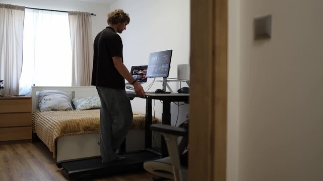 Young man raises an electric adjustable desk while standing on a walking pad setting the workstation height for comfortable computer use and better ergonomic posture at home