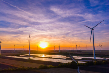 Obraz premium Wide aerial panorama of wind turbine farm and solar collectors at sunset. Colorful evening sky illuminates agricultural land filled with green power installations.