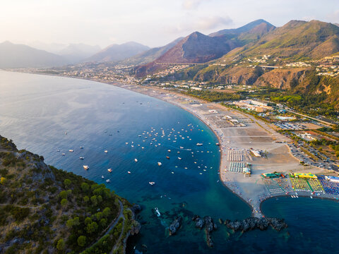 A breathtaking sunset aerial view captures Isola del Dino and the bay at Praia a Mare in Calabria, Italy. Boats navigate the deep blue waters along a sprawling beach, framed by dramatic coastal