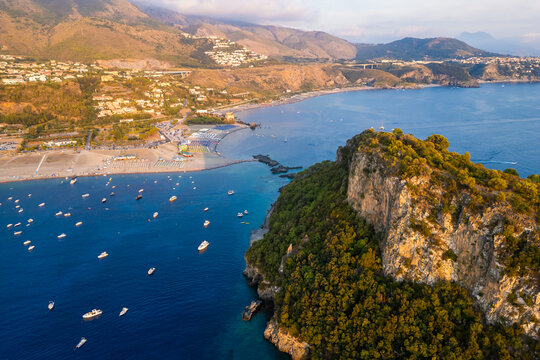 A breathtaking sunset aerial view captures Isola del Dino and the bay at Praia a Mare in Calabria, Italy. Boats navigate the deep blue waters along a sprawling beach, framed by dramatic coastal