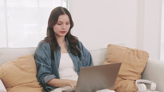 Tired young Asian woman closing her laptop, rubbing her eyes, and stretching after working from home.