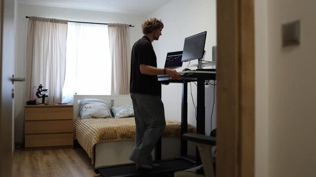 Young man walks on a treadmill while raising an electric standing desk to a comfortable height adjusting his computer workspace for active work and better posture at home