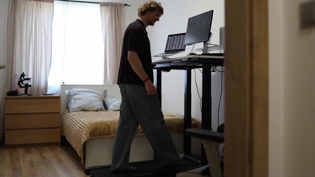 Young man walks on a treadmill while monitoring financial charts on computer screens turning screen time into an active routine that supports movement during remote work daily