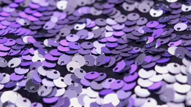 Close-up rotating shot of shimmering silver and purple sequins on black fabric background, highlighting texture and light reflection