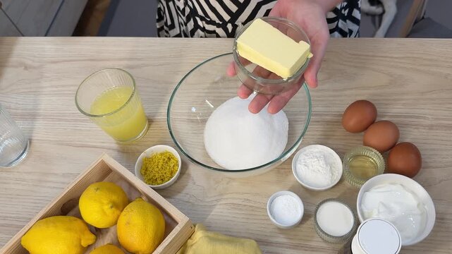 A wooden table displays baking ingredients arranged around a large clear glass bowl filled with white powder, alongside eggs, butter, lemons, and measuring cups