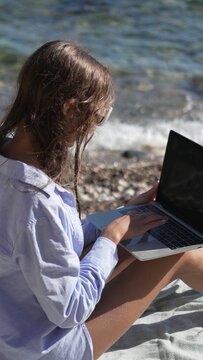 Remote work, beach, laptop. Young woman remotely working on her computer by the sparkling sea, enjoying a freelance lifestyle during summer vacation.