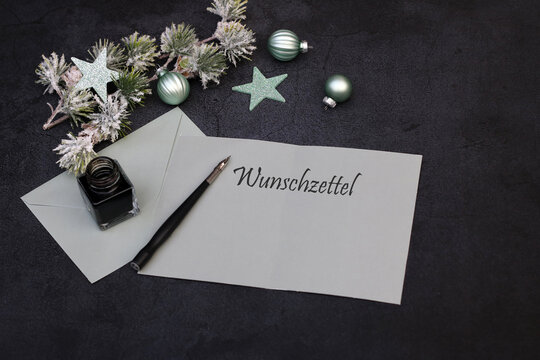 Mockup einer Weihnachtswunschliste mit dem deutschen Schriftzug &bdquo;Wunschzettel&ldquo;, salbeigr&uuml;nem Papier, Briefumschlag, F&uuml;llfederhalter, Tintenfass und gr&uuml;nen Christbaumkugeln auf dunklem Hintergrund.