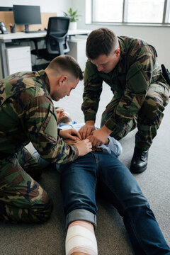 Military personnel perform emergency medical training on man lying on floor. Soldiers practice life saving first aid skills in office. Team provides urgent care, CPR simulation for injured person