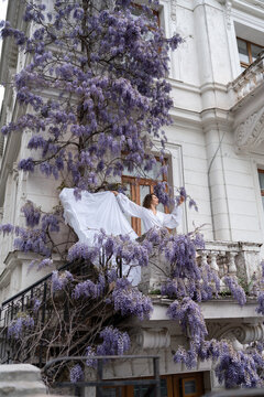 Elegant female in white dress posing on historical building balcony adorned with purple wisteria flowers during spring season