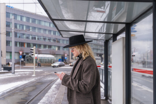 Young man waiting at tram stop using smartphone in urban winter setting