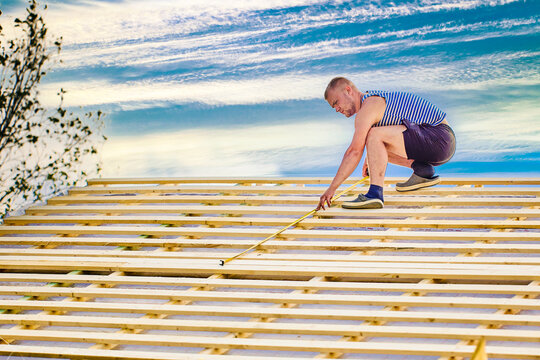 Measuring roof battens with tape measure while checking spacing and alignment, worker prepares wooden roof structure for installation of roofing materials.