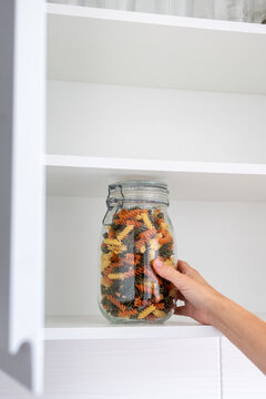 Hand taking a jar of colorful pasta from an organized kitchen pantry