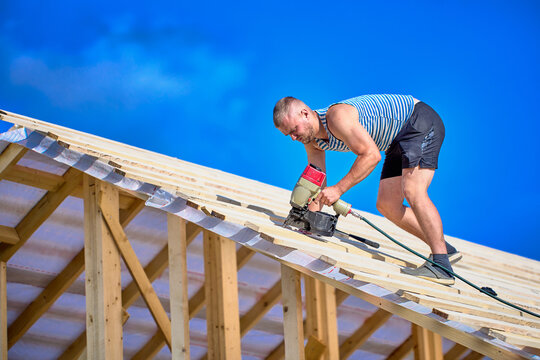 Securing timber battens over roofing underlay, worker uses nail gun while assembling wooden roof structure during construction.