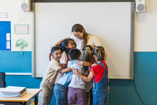 Happy teacher smiling while embracing a group of diverse elementary school students in a classroom, showing support and community