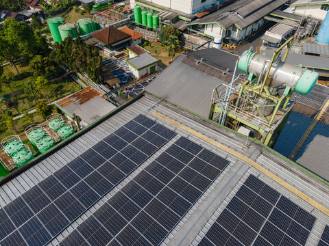 Aerial view of solar panels installed on the roof of an industrial facility with green storage tanks and surrounding trees in Banyumas Regency, Central Java, Indonesia.