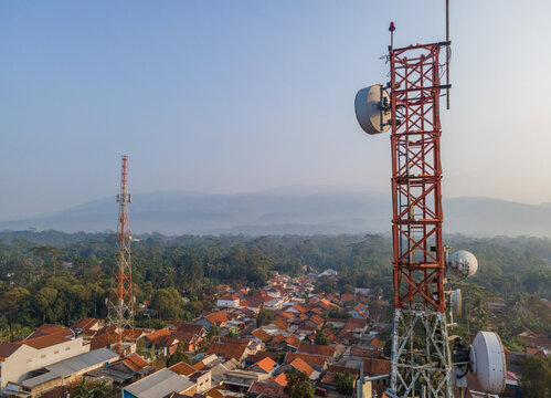 Aerial view of telecommunication towers with satellite dishes overlooking a residential area with tiled rooftops and lush trees in the morning light in Cilongok, Central Java, Indonesia.