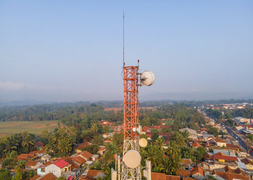 Aerial view of a telecommunications tower rising above a residential neighborhood with lush palm trees and tiled rooftops in Cilongok, Central Java, Indonesia.